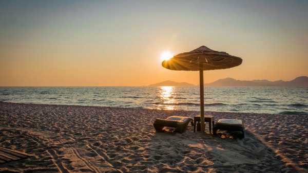 Beach scene at sunset with two lounge chairs and a straw umbrella facing calm ocean water, sunlight reflecting on waves and distant mountains on the horizon.