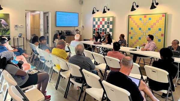 Community meeting in a bright room; attendees sit in rows facing a panel at tables, with a presentation screen and two large board displays on the wall behind the speakers.